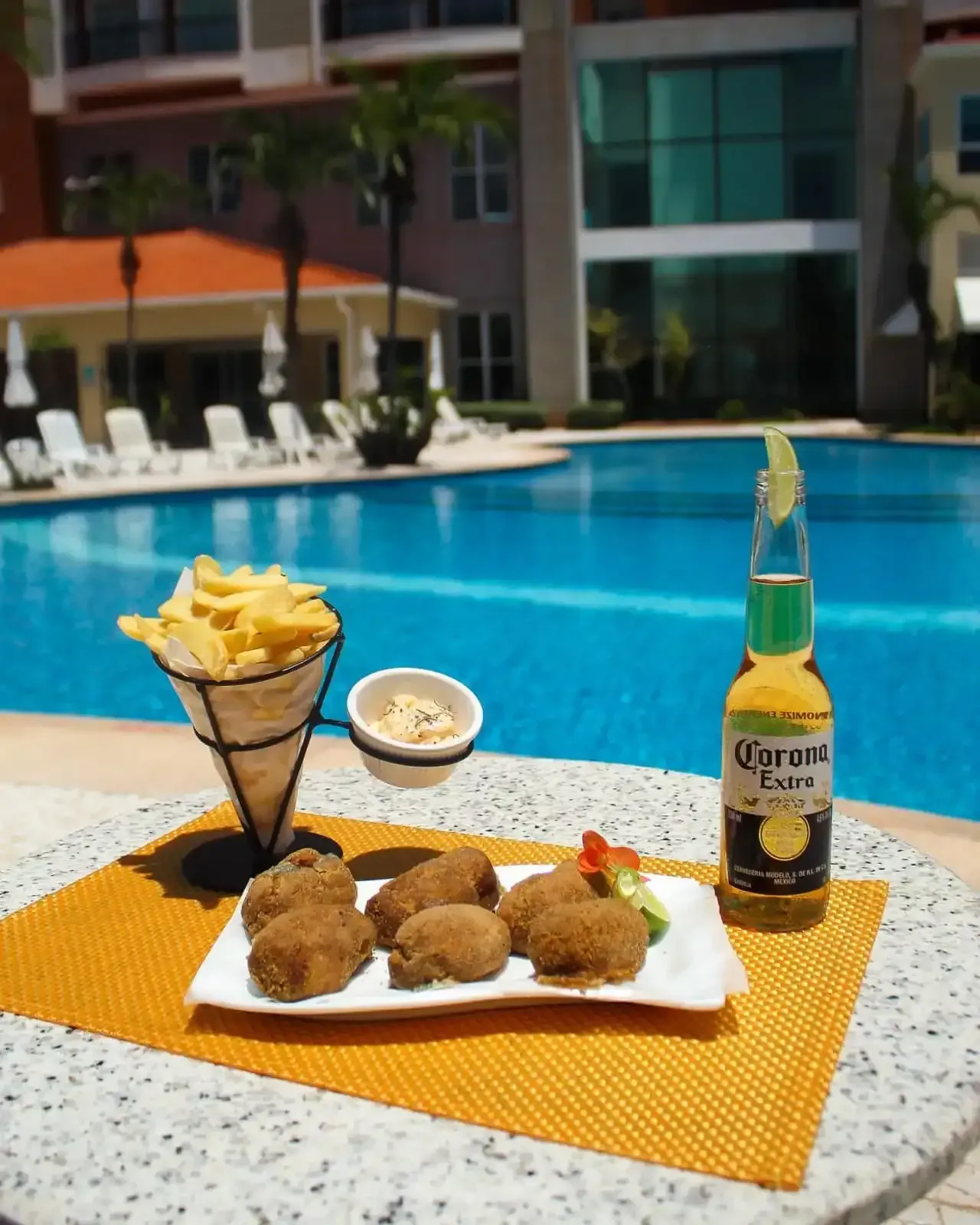 Petiscos à beira da piscina: batatas fritas, bolinhos fritos, molho, garrafa de cerveja Corona com fatia de limão. Dia ensolarado.
