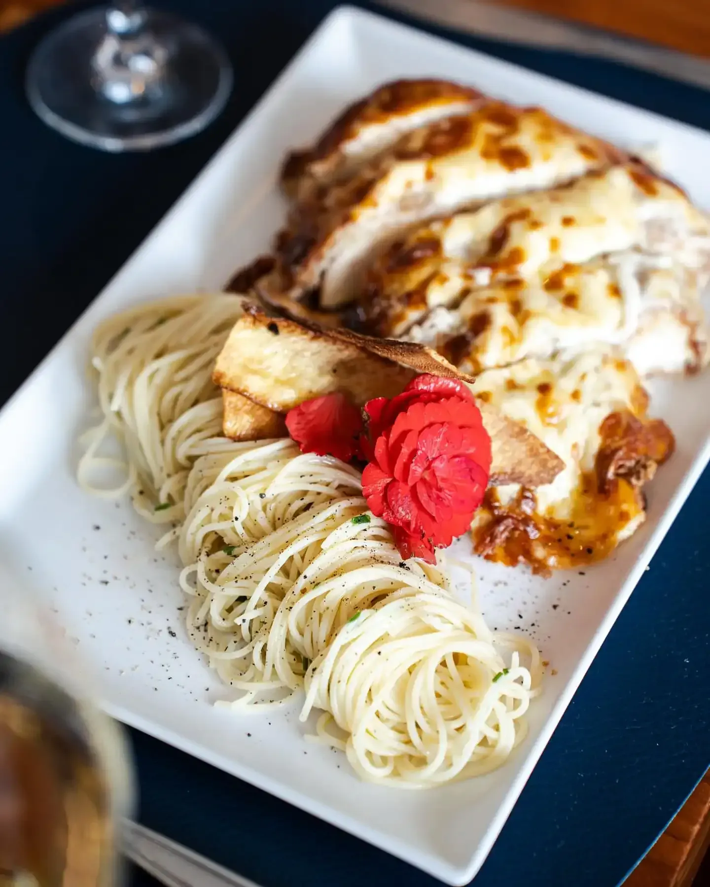 Prato de frango com massa em um prato branco; o frango é coberto com queijo e a massa está ao lado; uma guarnição vermelha decora a massa.