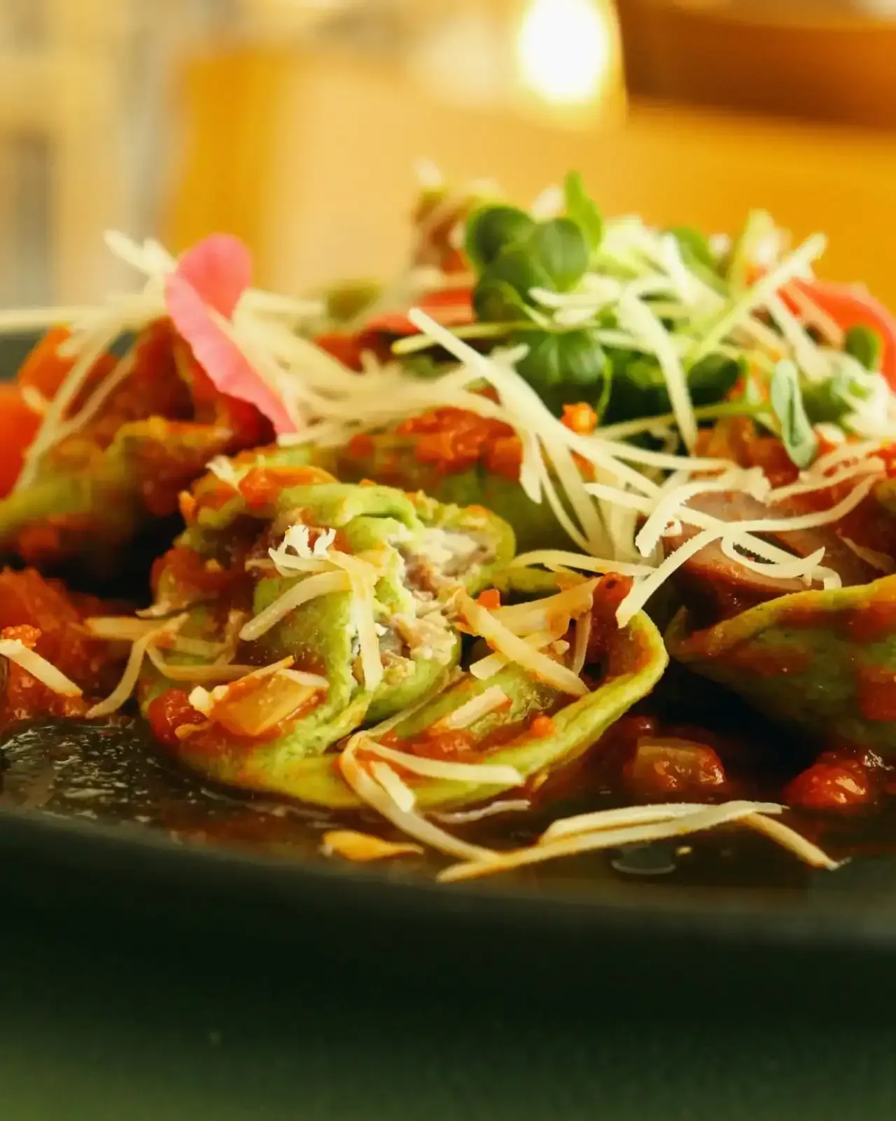 Ravioli verde com molho vermelho, queijo e microverdes em um prato escuro.