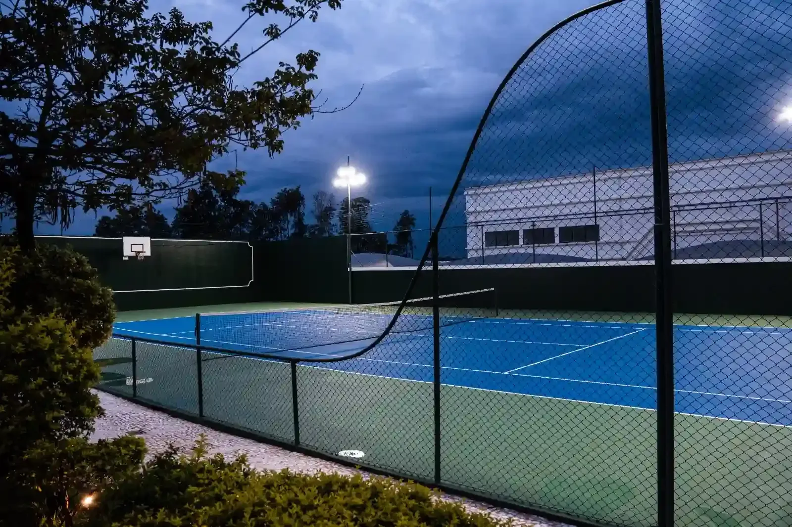 Quadra esportiva azul e verde ao entardecer, iluminada por luzes suspensas, com uma cerca e uma árvore em primeiro plano.