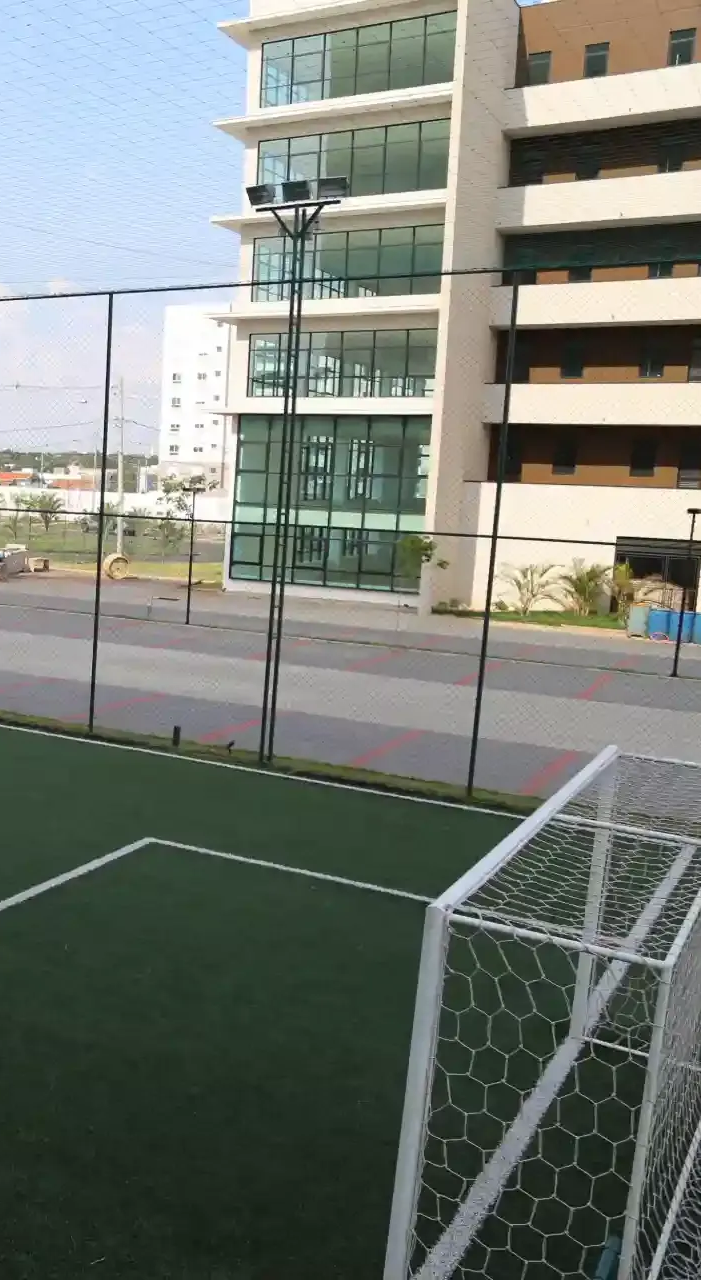 Campo de futebol com gol em primeiro plano e prédios ao fundo. Grama verde e cerca preta.