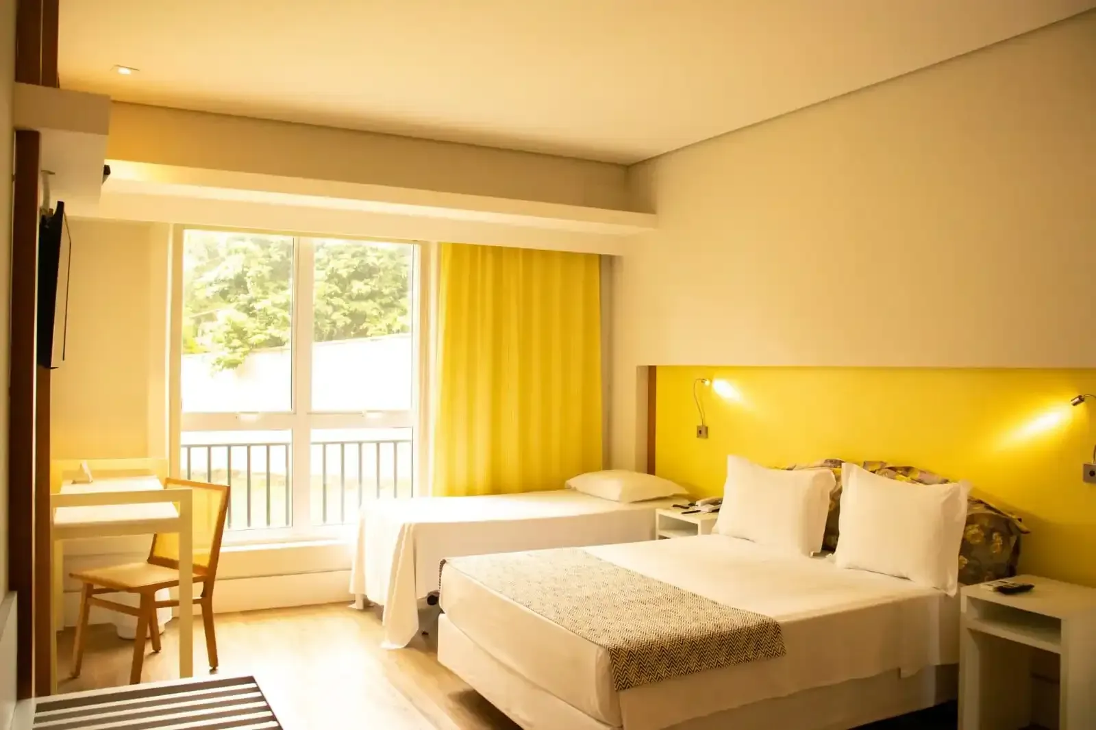 Quarto de hotel com duas camas, uma escrivaninha e uma janela com cortinas amarelas.