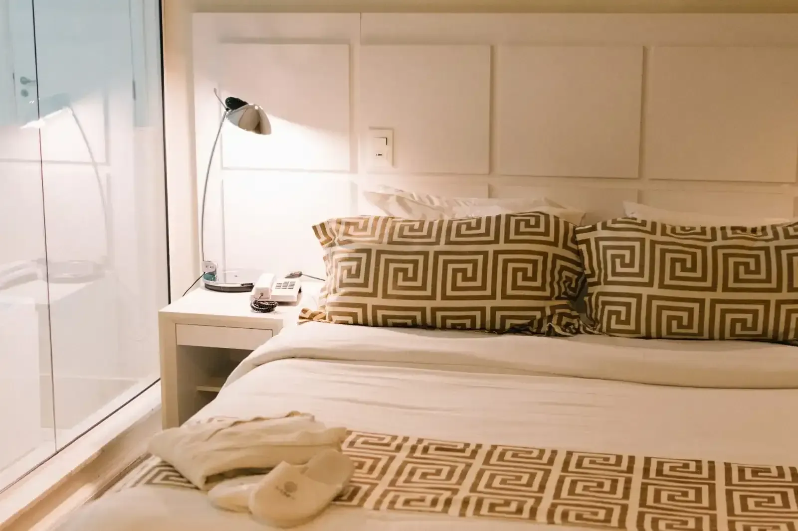 Quarto com cabeceira branca, cama, mesa de cabeceira, abajur, almofadas estampadas e peseira.