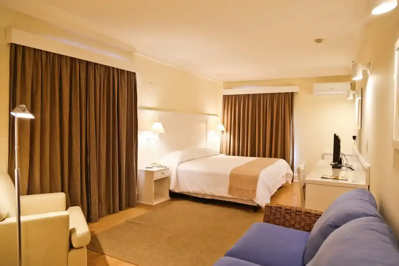 Quarto de hotel com cama, sofá e televisão. Cortinas marrons e paredes bege.