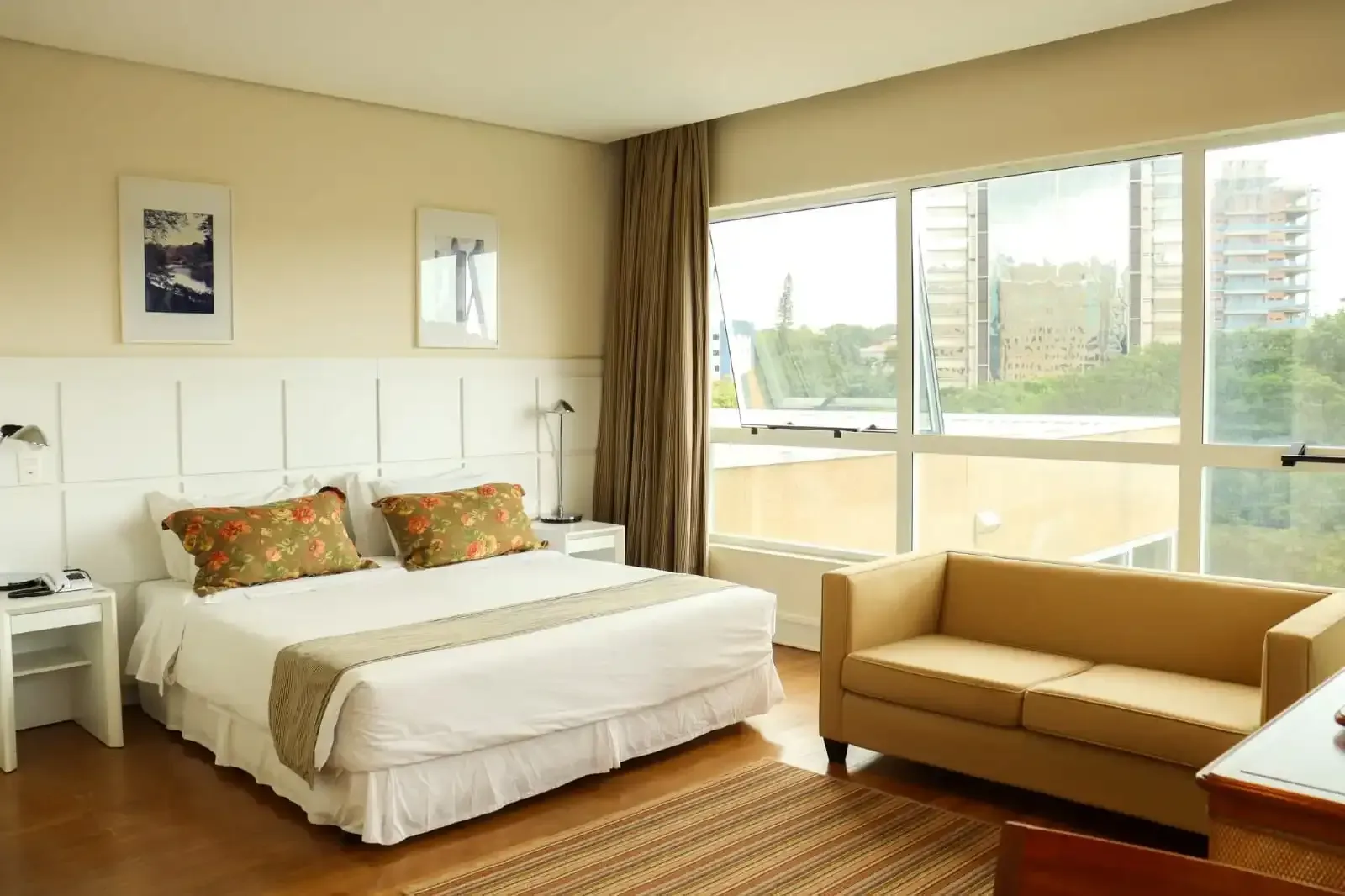 Quarto de hotel com cama, sofá pequeno e janelas amplas com vista para os prédios da cidade.