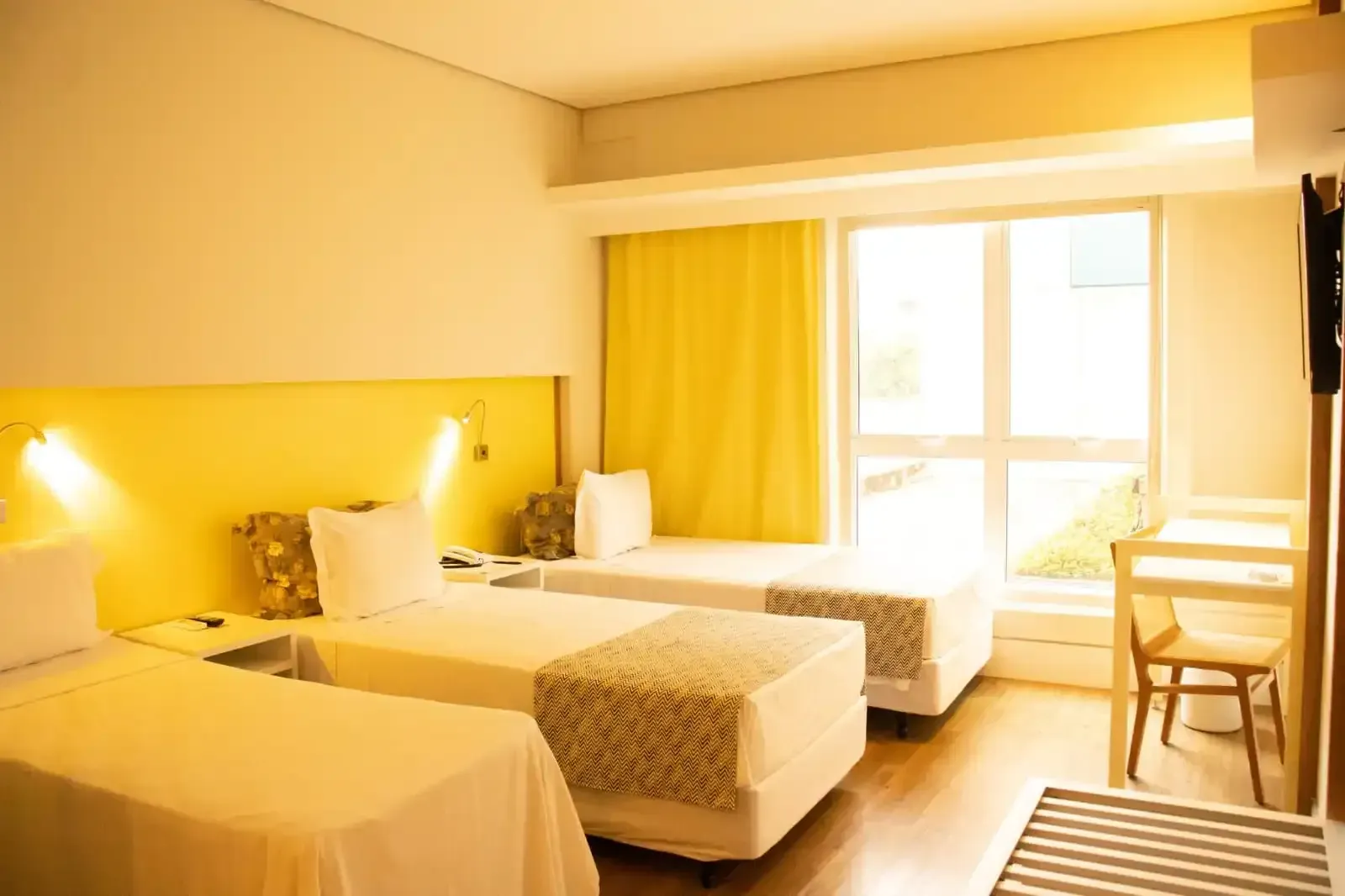 Um quarto de hotel bem iluminado com três camas de solteiro, uma escrivaninha e uma janela com detalhes em amarelo.