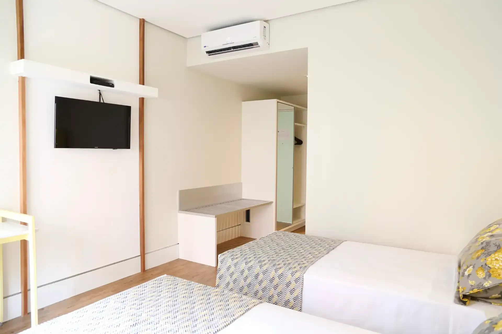 Quarto com duas camas de solteiro, escrivaninha pequena, armário e TV de parede.