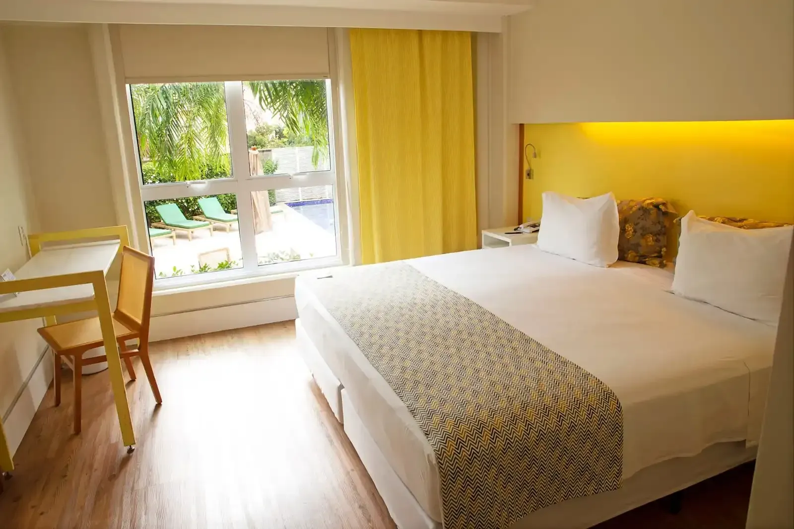 Quarto de hotel com cama, escrivaninha e janela com vista para a área da piscina. Detalhes em amarelo.