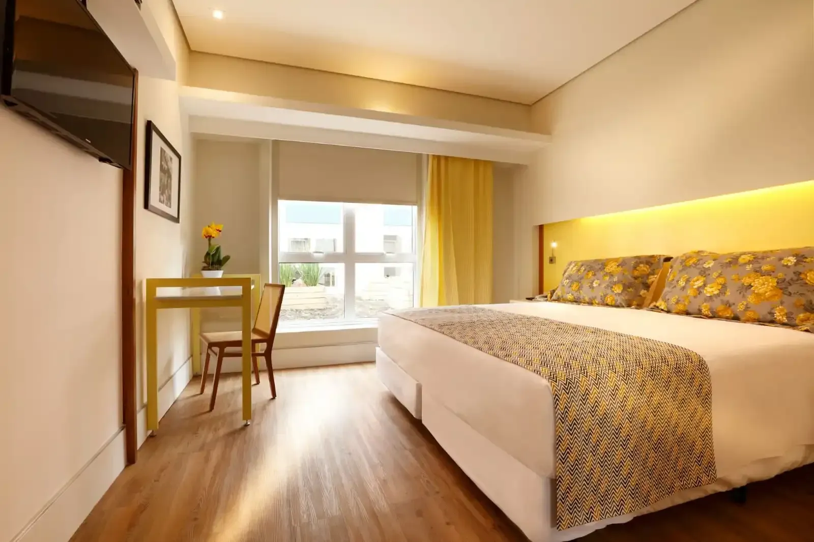 Quarto de hotel com cama de casal, escrivaninha e janela grande com cortinas amarelo-claras.