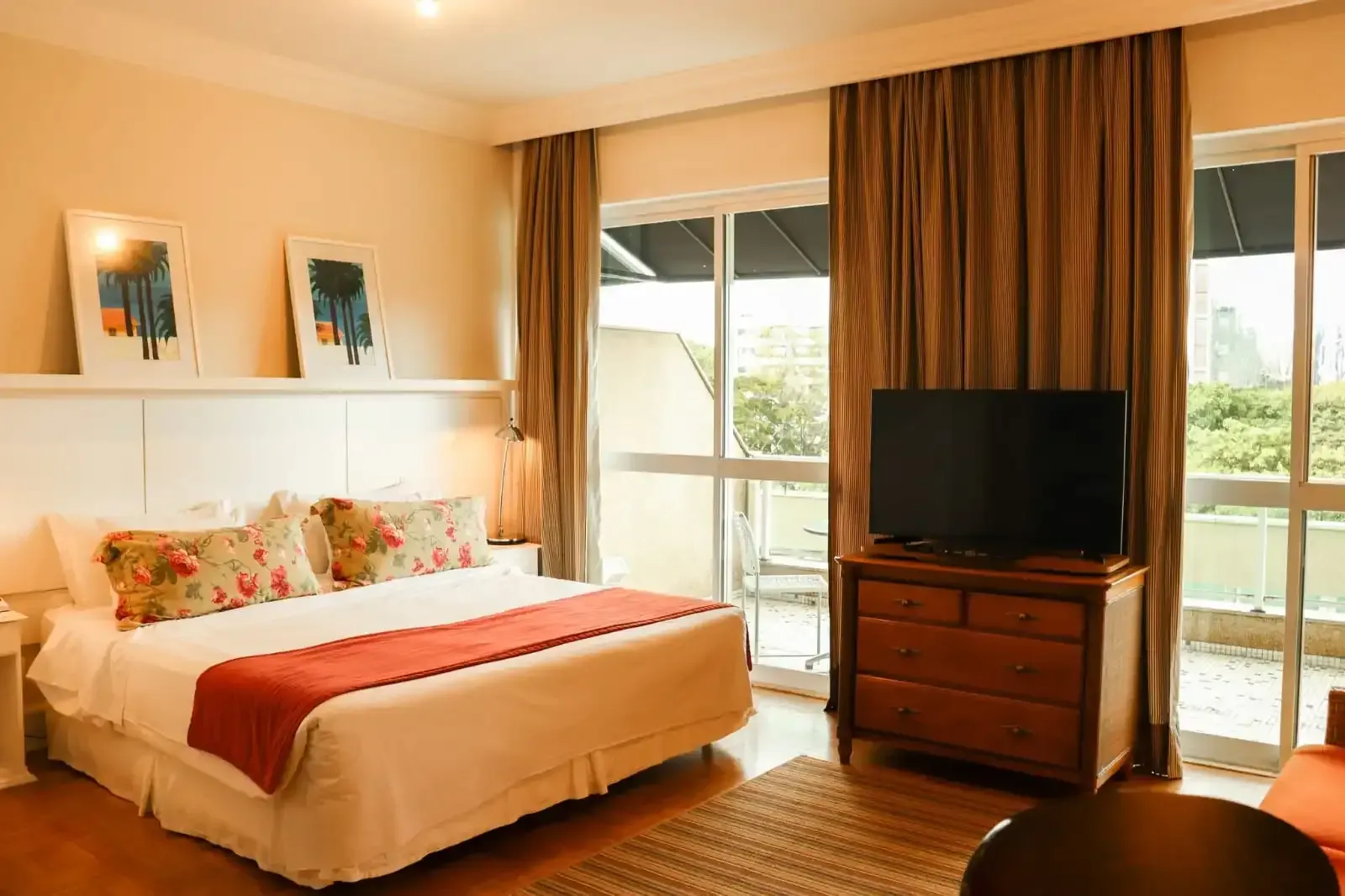 Quarto com cama, TV e acesso à varanda. Paredes bege, cortinas marrons e móveis em tons neutros.