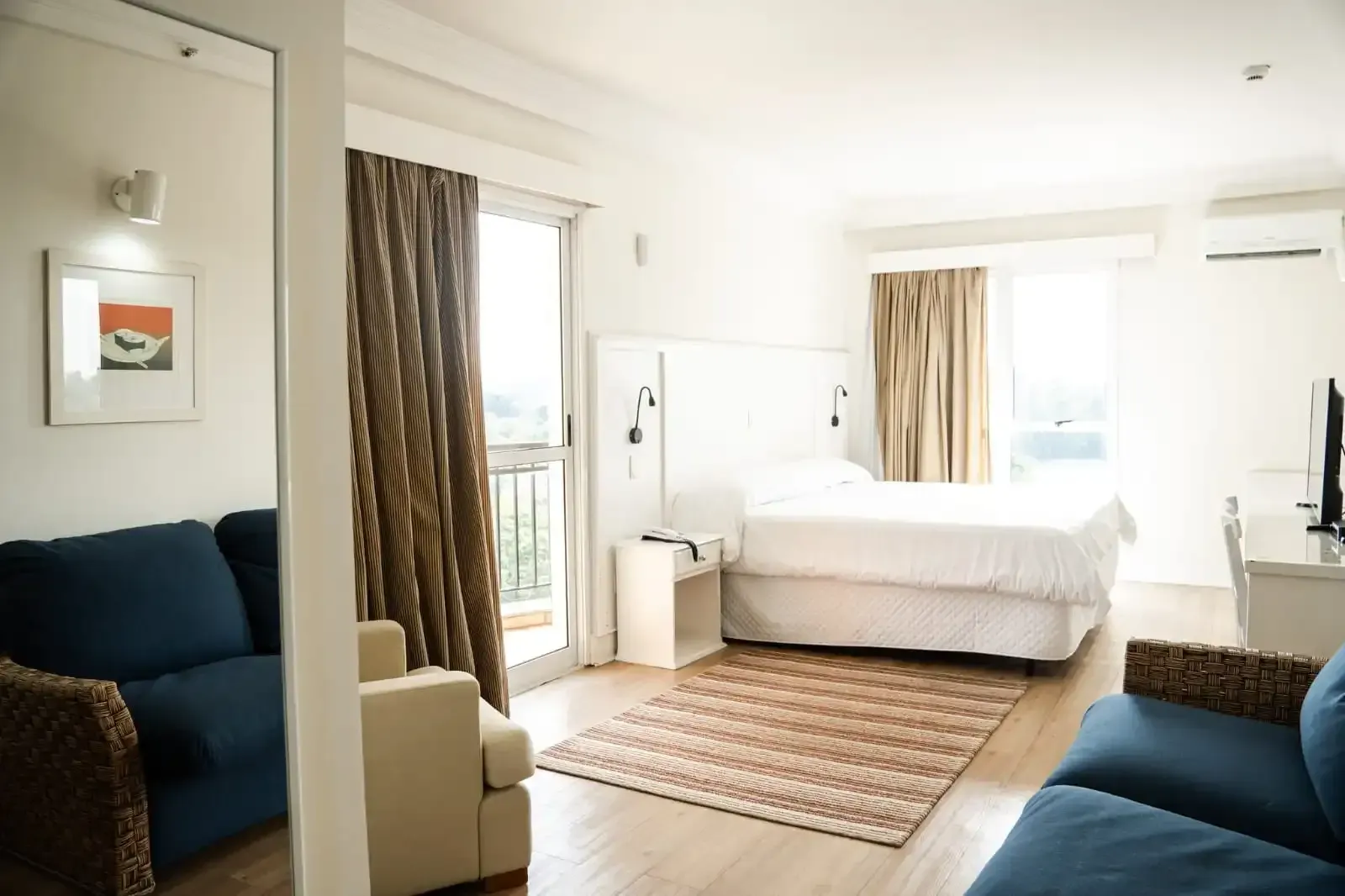 Quarto de hotel luminoso com cama, área de estar e acesso à varanda.
