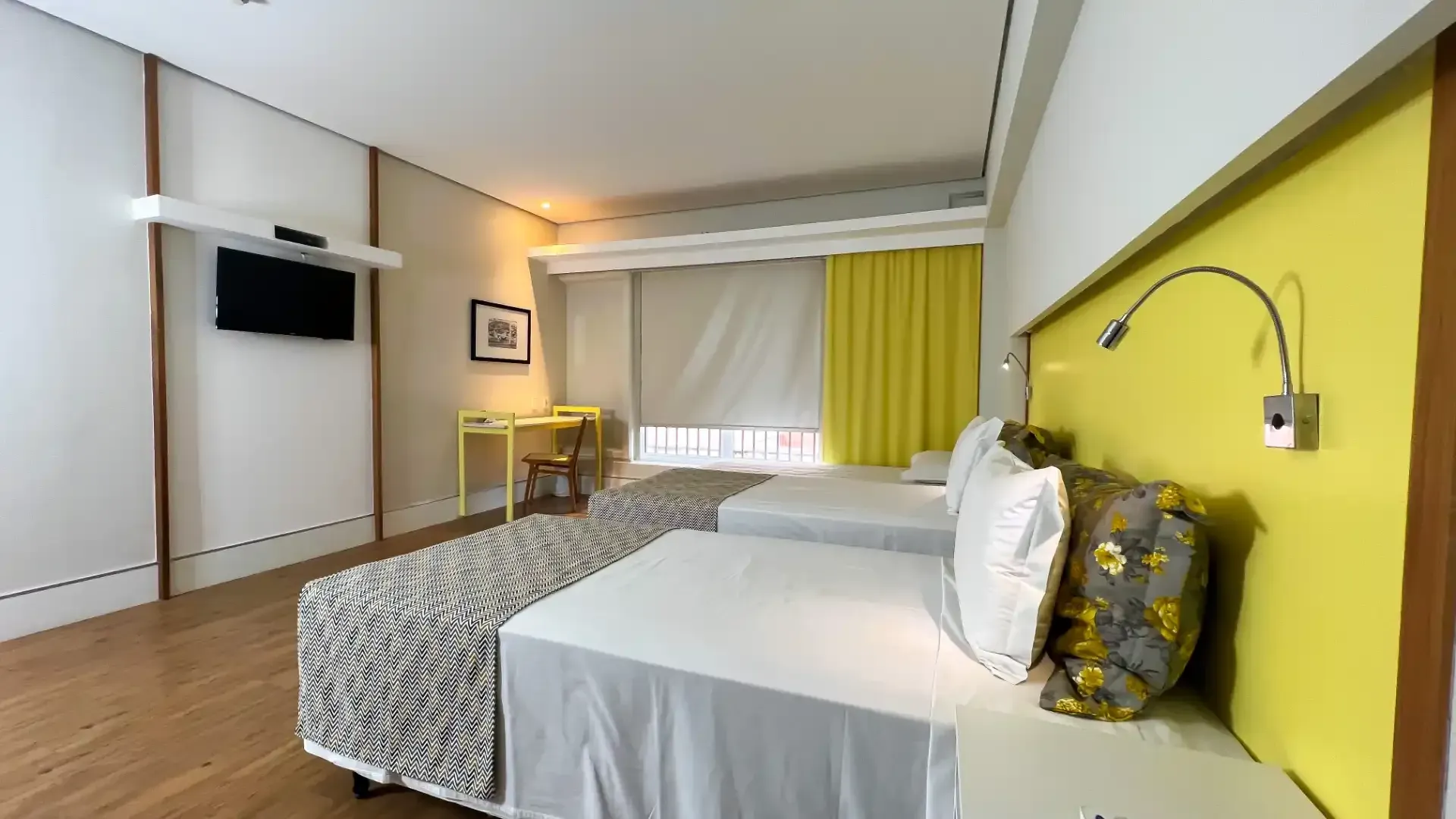 Duas camas em um quarto de hotel com lençóis brancos, detalhes em amarelo e uma televisão fixada na parede.