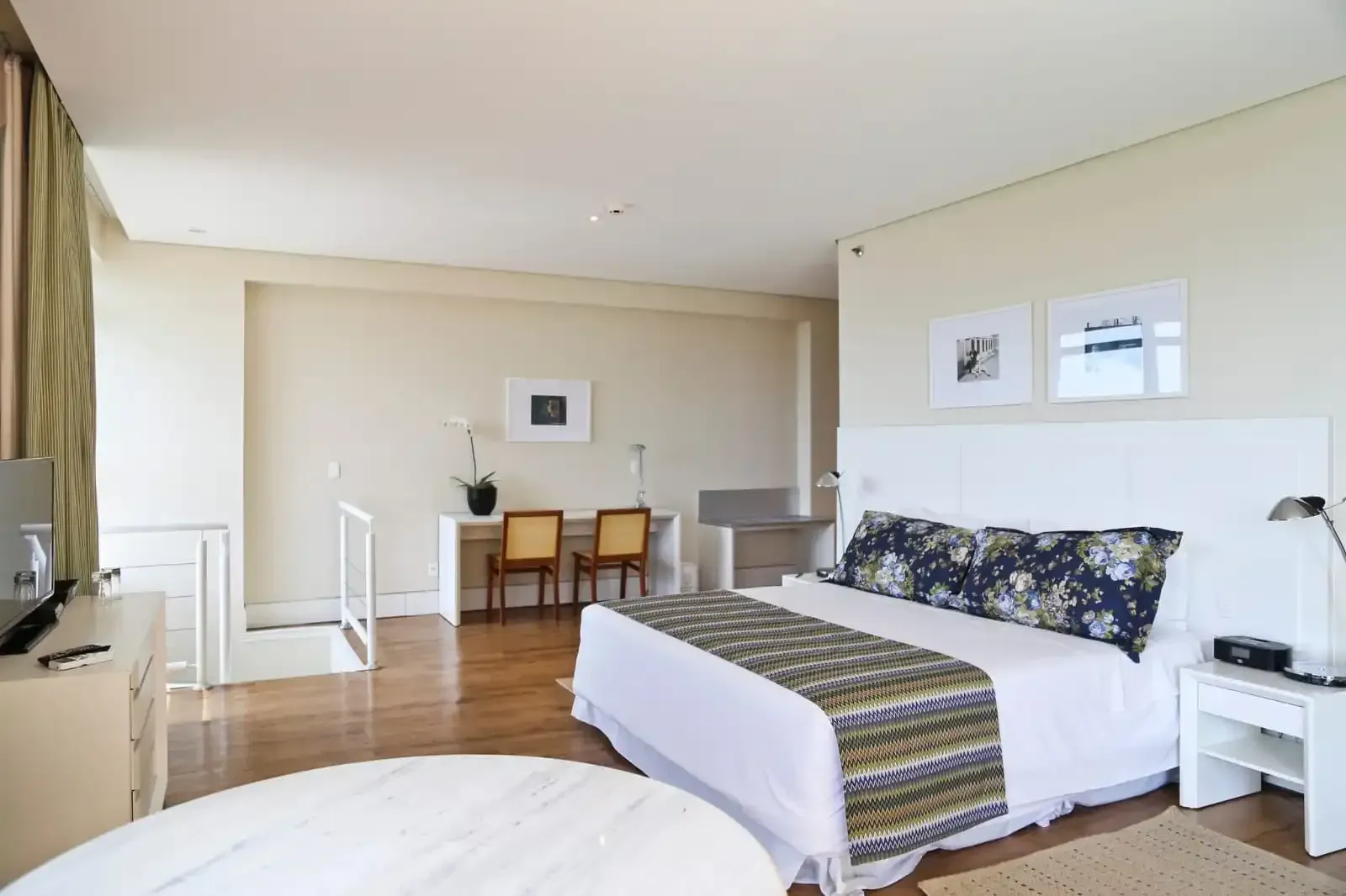 Quarto de hotel com cama, duas cadeiras e uma pequena mesa sobre piso de madeira.