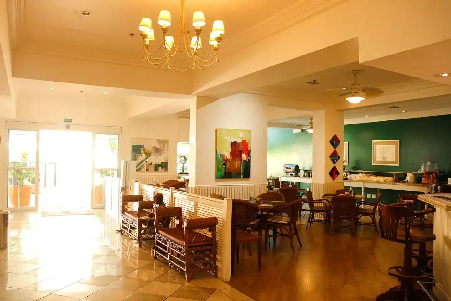Interior luminoso com mesas, cadeiras e obras de arte. A entrada com portas de vidro fica à esquerda.