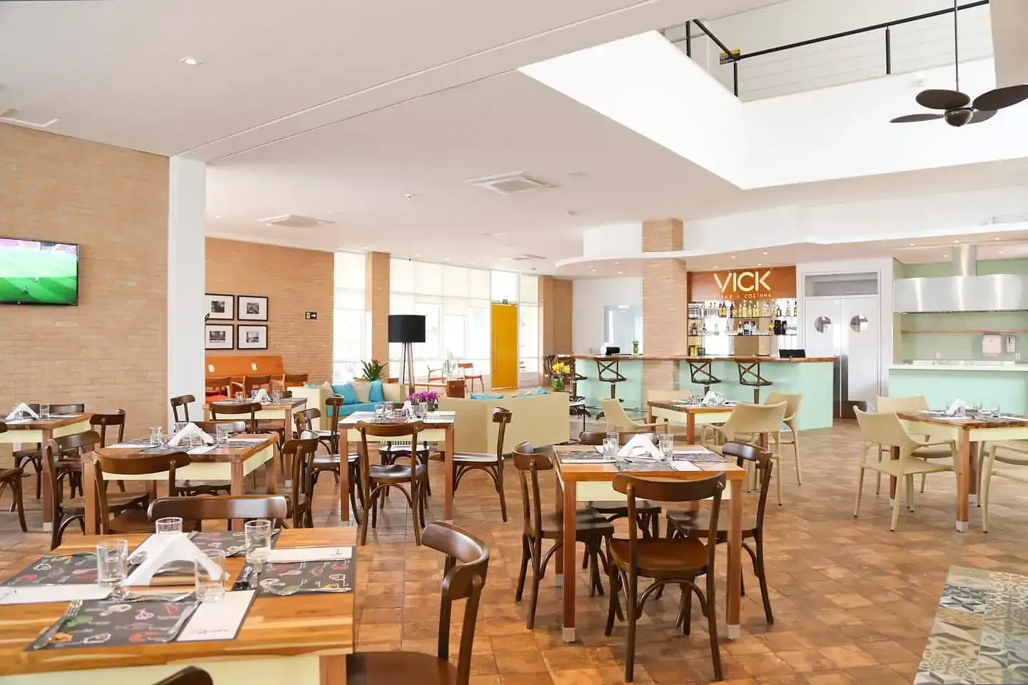 Interior de restaurante com mesas, cadeiras, bar e TV. Cores neutras, luz natural e ambiente descontraído.