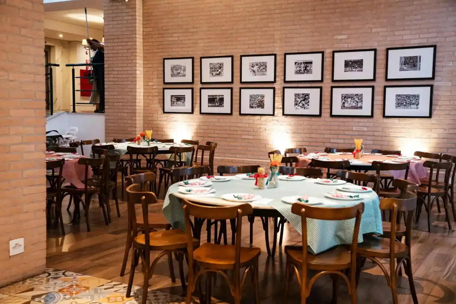Interior de restaurante com mesas redondas, cadeiras de madeira, paredes de tijolos aparentes, fotos emolduradas e toalhas de mesa coloridas.