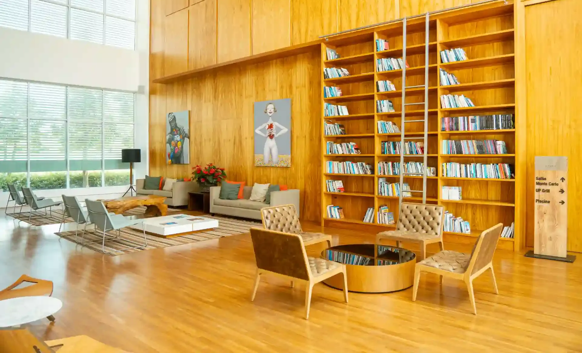 Interior da biblioteca com painéis de madeira, estantes de livros, assentos e grandes janelas.