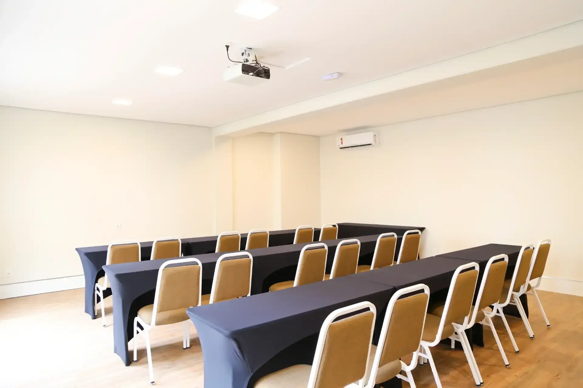 Sala de conferências com mesas em formato de U, cadeiras bege, projetor e aparelho de ar condicionado.
