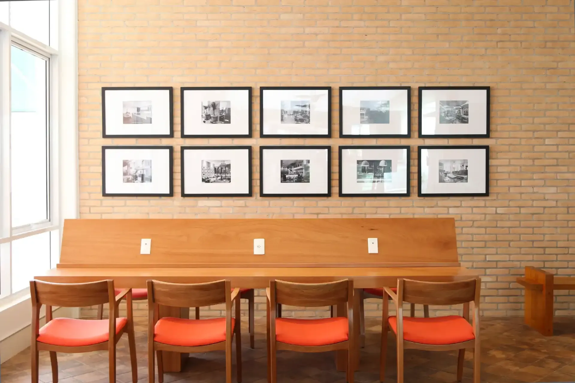 Uma fileira de fotos emolduradas acima de um banco de madeira com cadeiras laranjas encostado em uma parede de tijolos.