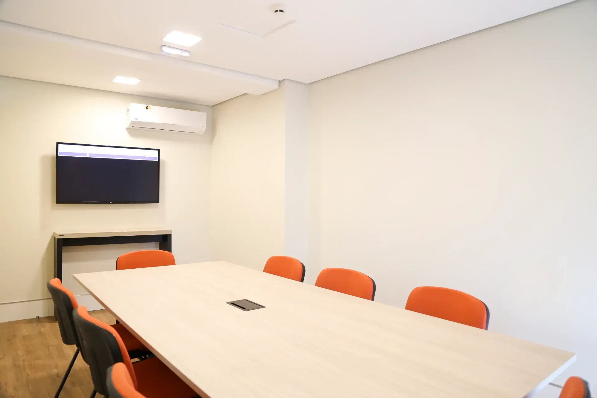 Sala de conferências com mesa comprida e cadeiras laranja, TV na parede.