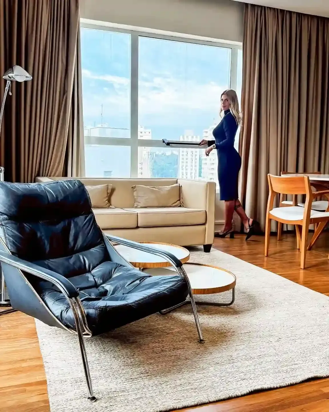 Mulher olhando pela janela em um apartamento moderno. Ela segura uma bandeja. O interior apresenta um sofá, uma cadeira e vista da cidade.