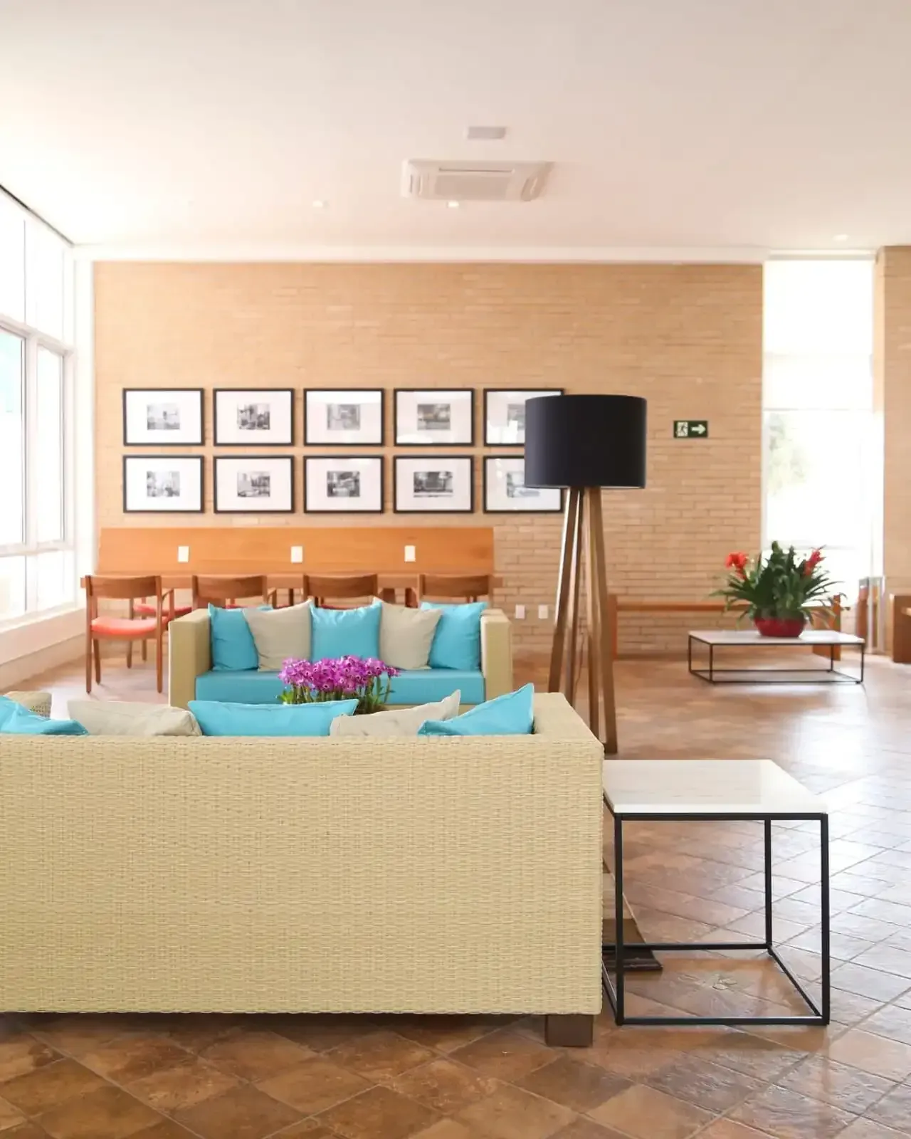 Uma sala de estar moderna com sofá bege, almofadas verde-azuladas, luminária de chão preta e fotos emolduradas em uma parede de tijolos cor de areia.