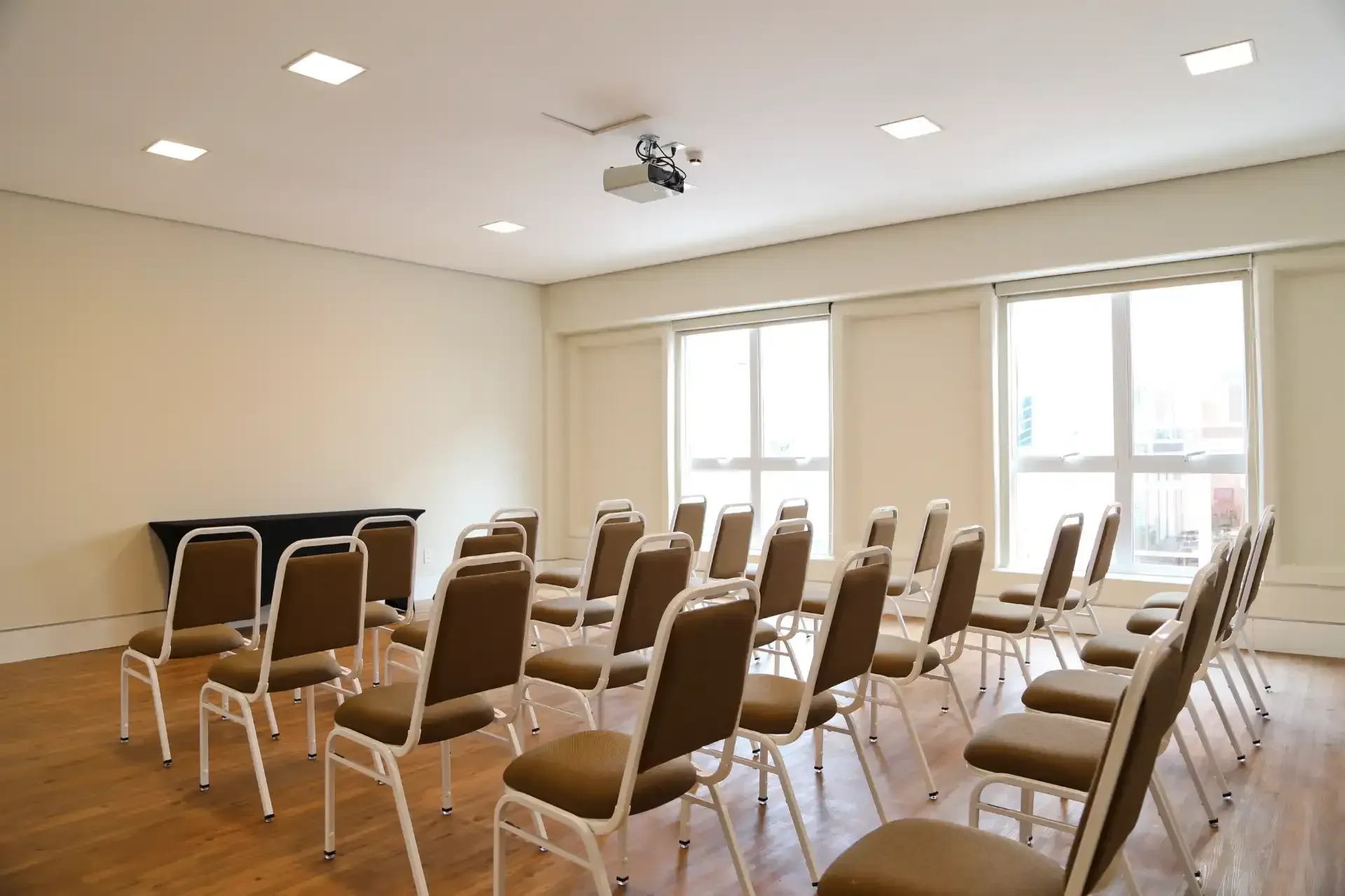 Sala de reuniões vazia com fileiras de cadeiras marrons, janelas e projetor.
