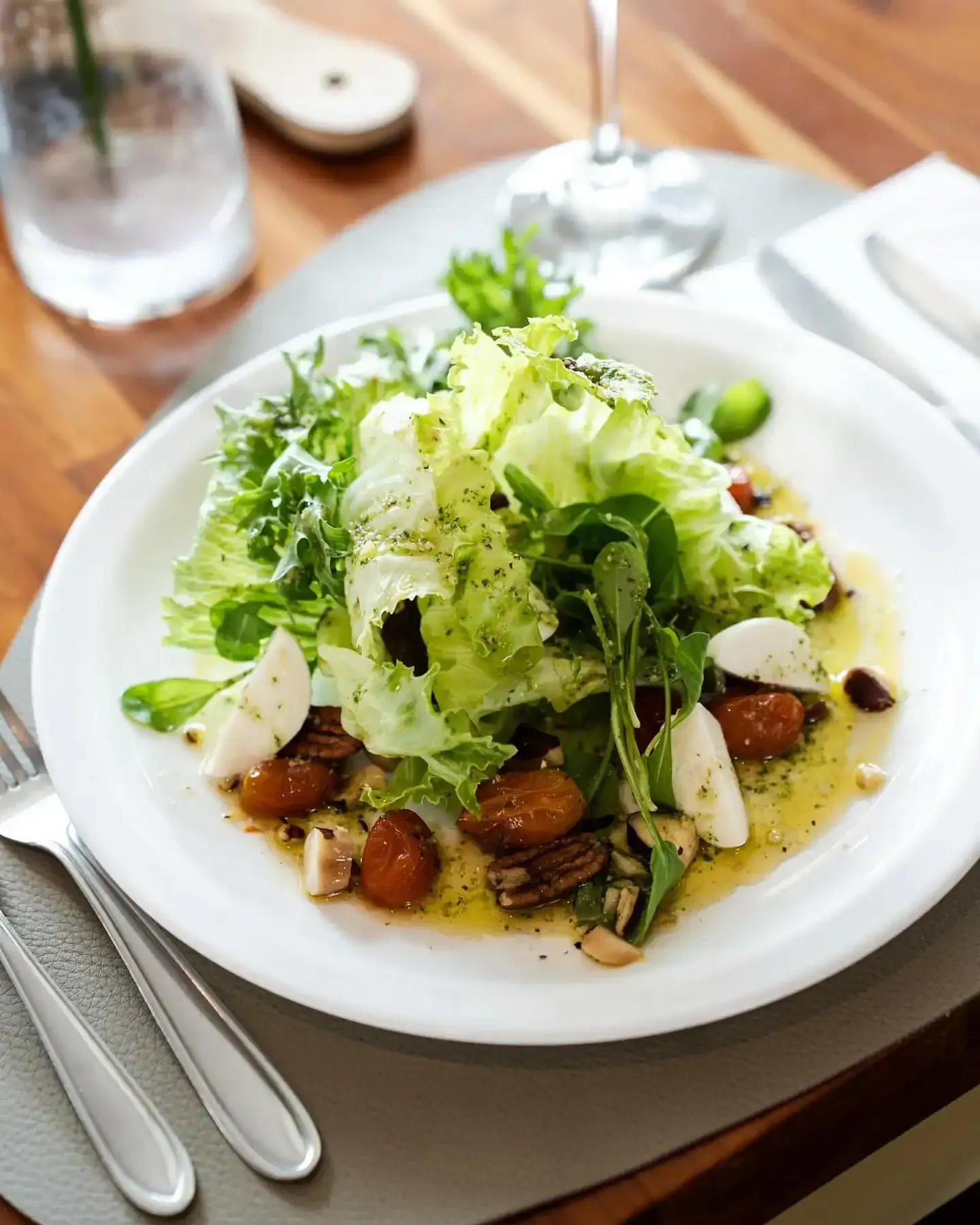 Salada com folhas verdes, nozes e tomates em um prato branco, servida com talheres e copo de água sobre uma mesa de madeira.