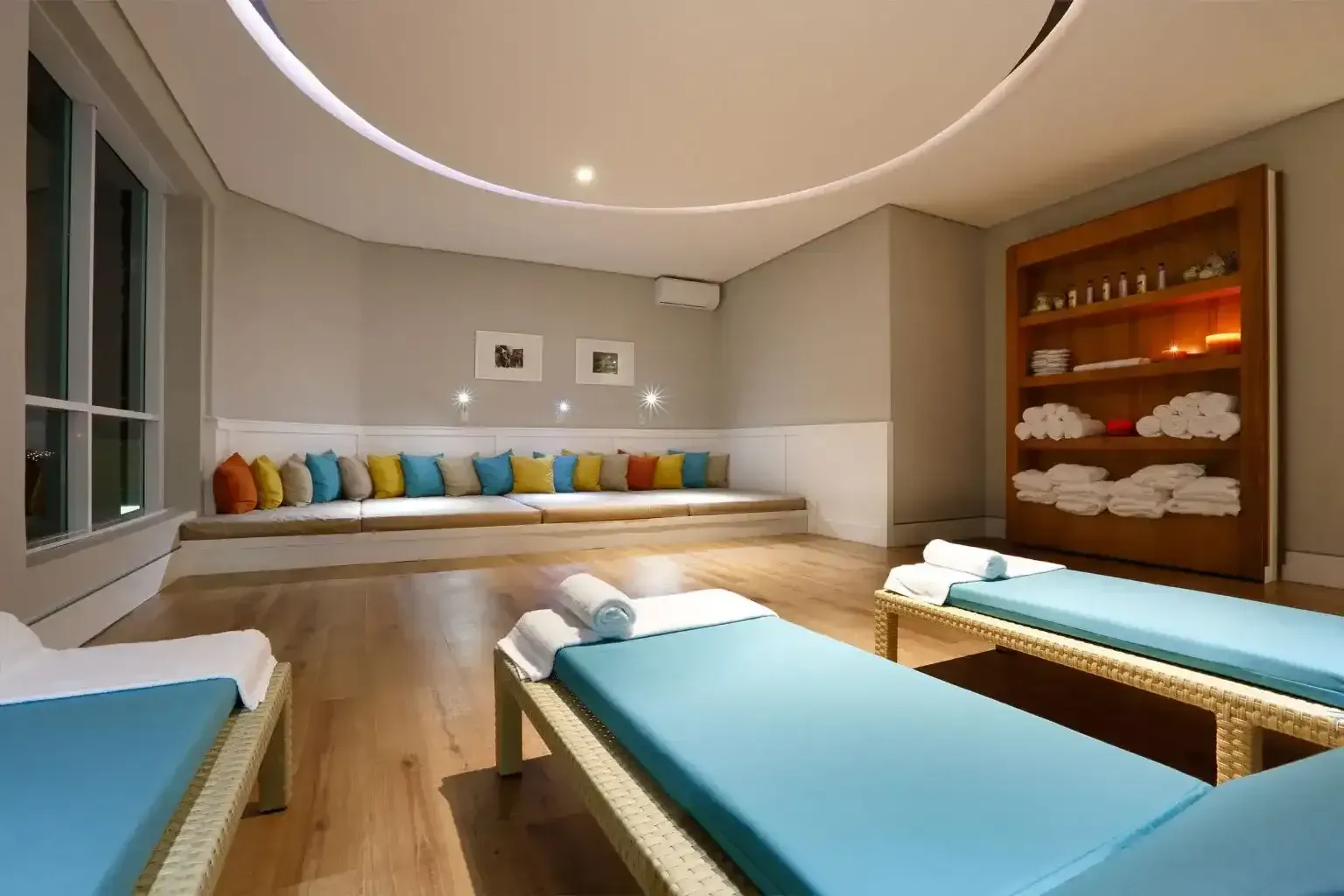 Sala de spa com camas de massagem, banco almofadado, prateleira de madeira com toalhas e almofadas coloridas.