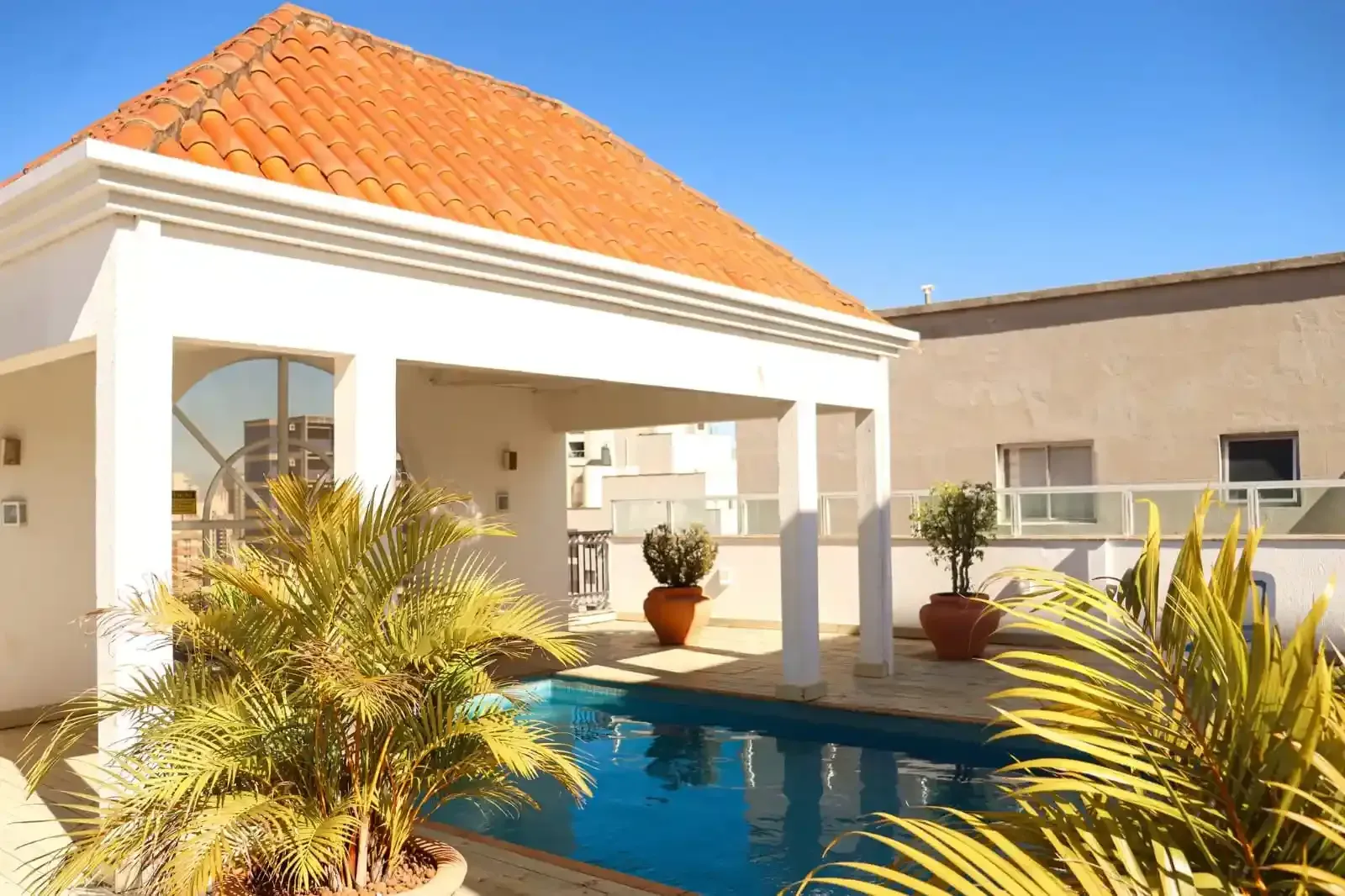 Piscina na cobertura com estrutura branca e telhado de telhas laranja, rodeada de plantas e sob um céu ensolarado.