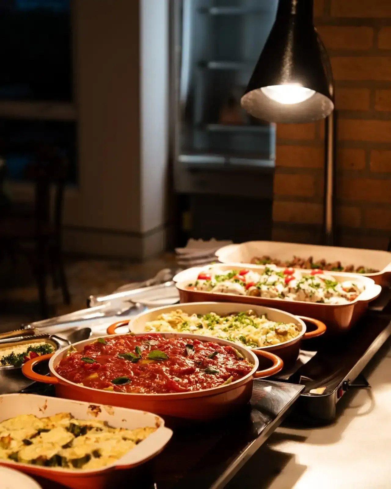 Buffet com diversos pratos servidos em travessas de terracota sob uma lâmpada aquecedora.