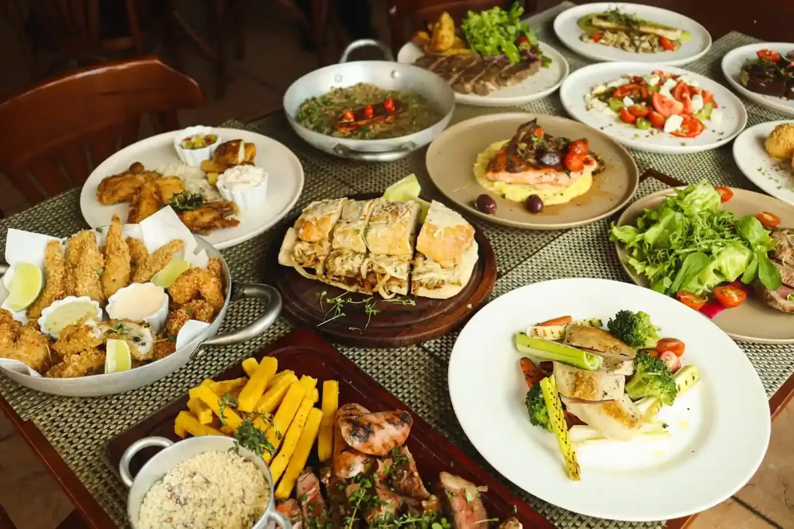 Mesa repleta de diversos pratos: bife, peixe, pão, saladas e acompanhamentos.
