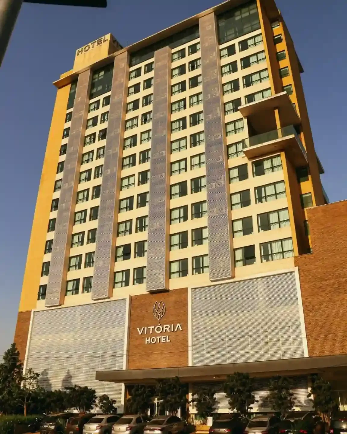 Exterior do Hotel Vitória: Prédio alto e moderno com fachada, janelas e sinalização em tons de amarelo e marrom. Carros estacionados embaixo.