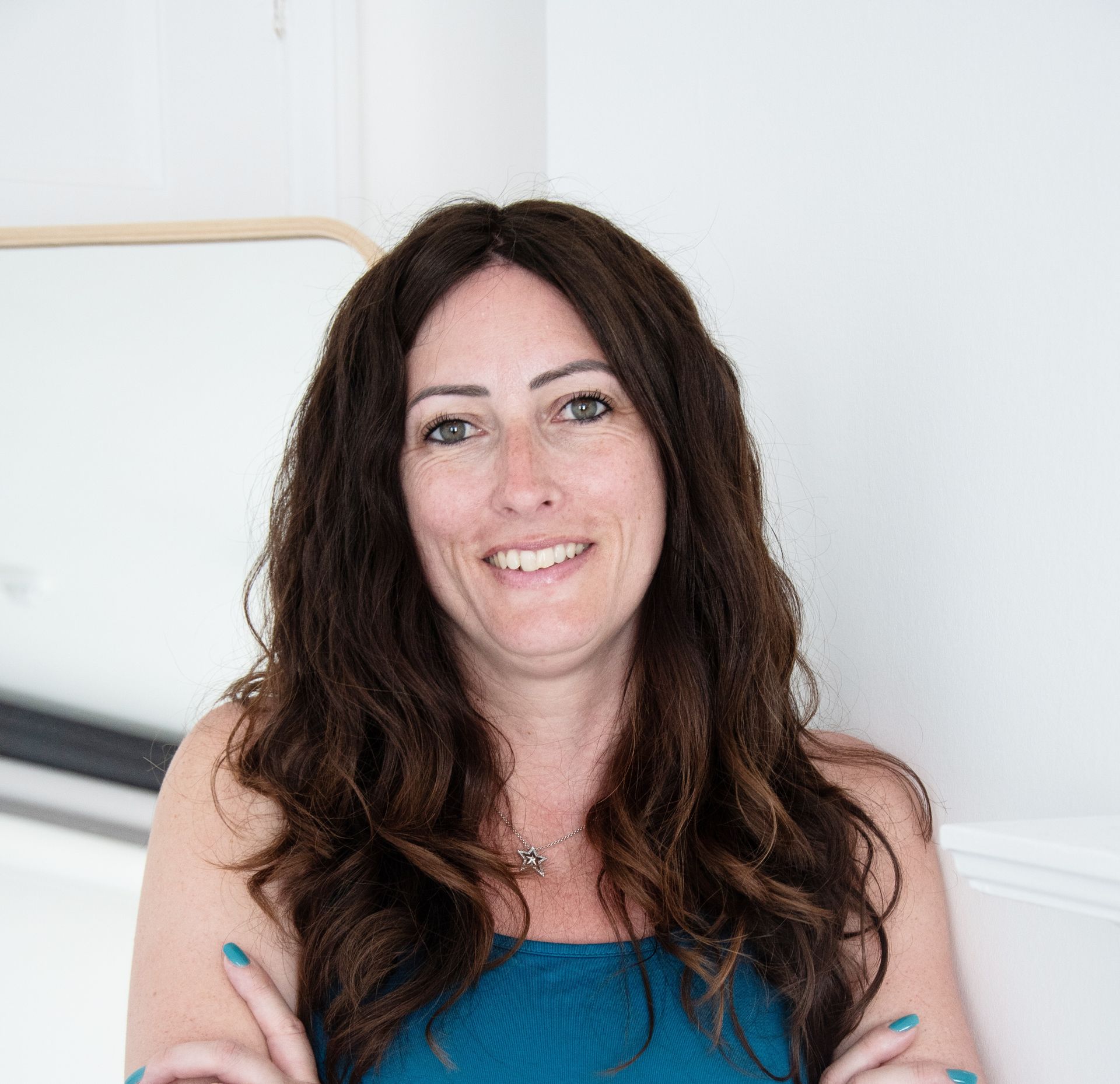 Woman smiling, holding cup, working on laptop at white desk. Blue floral robe, plant and window background.