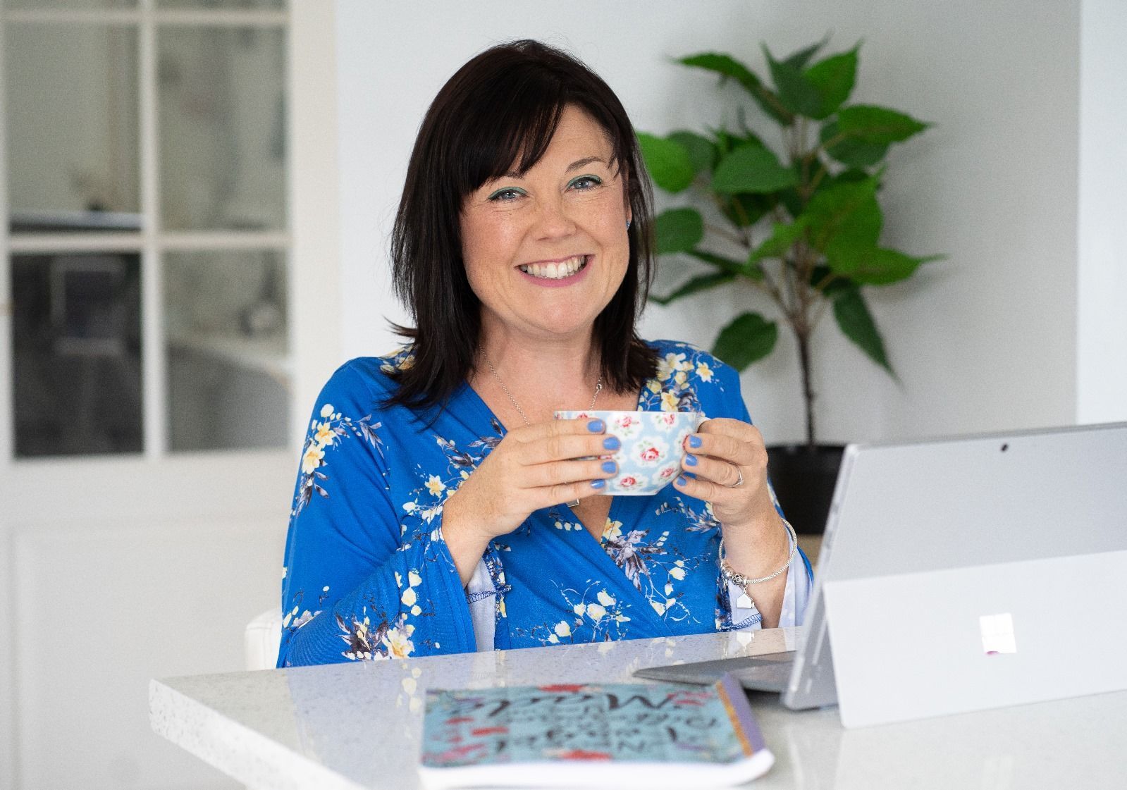Woman smiling, holding cup, working on laptop at white desk. Blue floral robe, plant and window background.