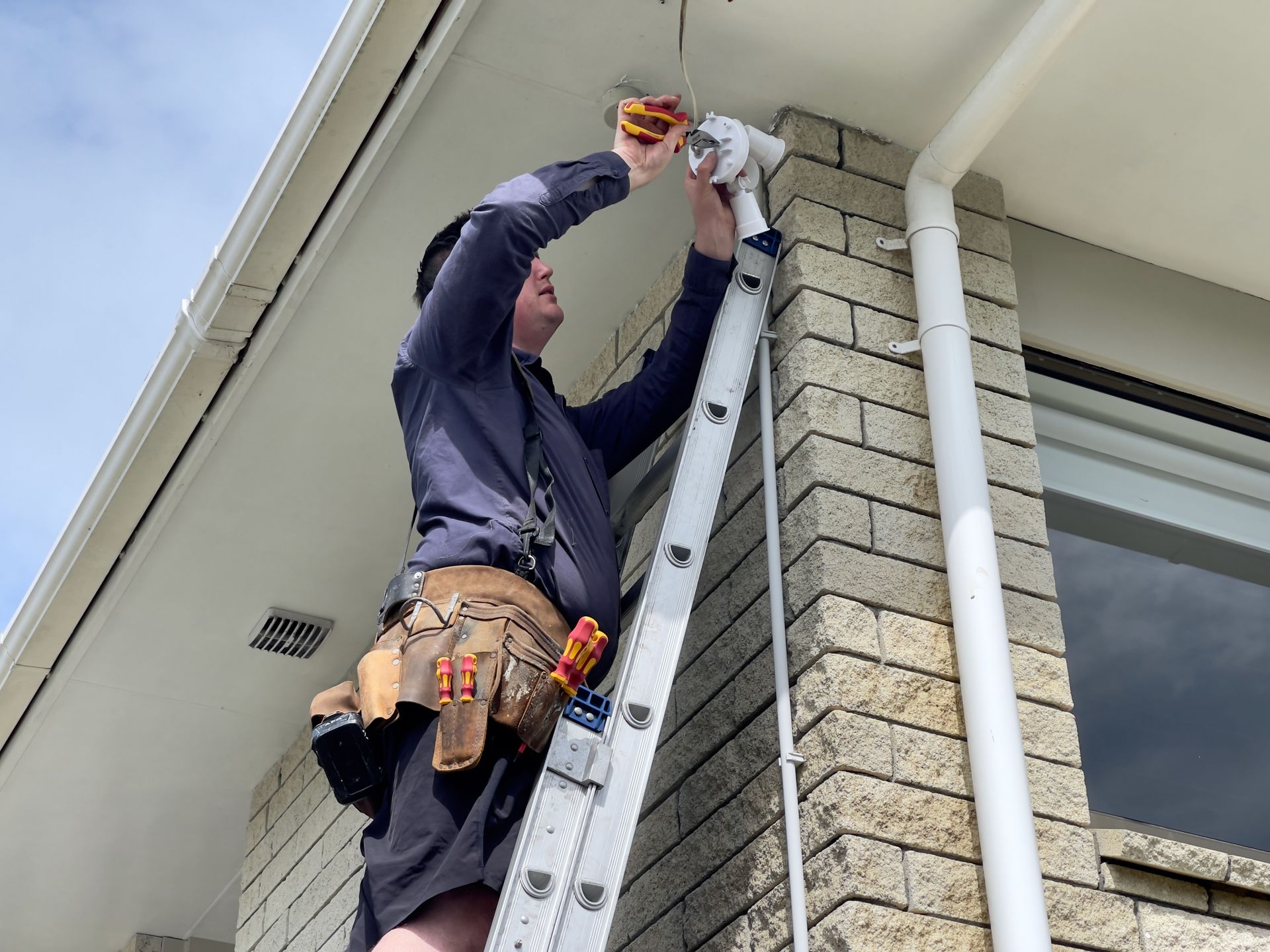 Electrician on a ladder installing wiring on a brick building exterior.