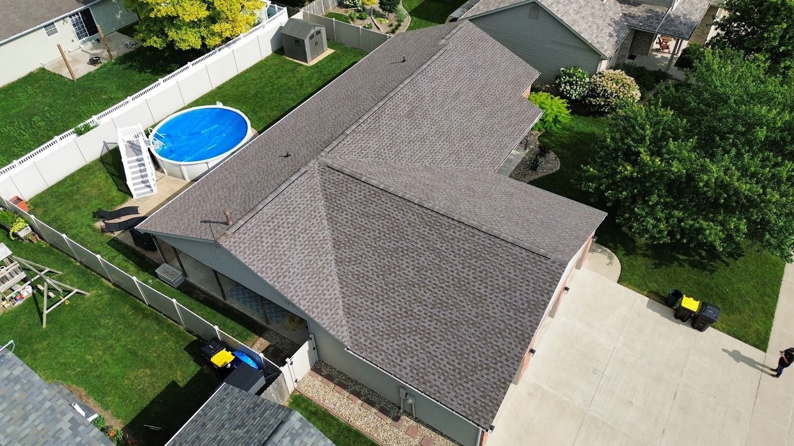 Overhead view of a house with a brown shingled roof, surrounded by green grass and trees in a sunny setting.