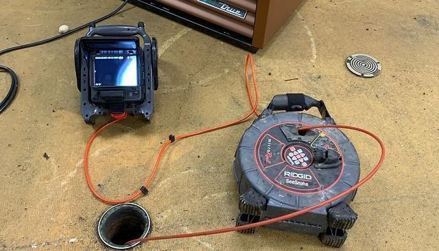 Camera inspecting a pipe: equipment on dirt, orange cable, monitor shows inside the pipe, by wooden structure.