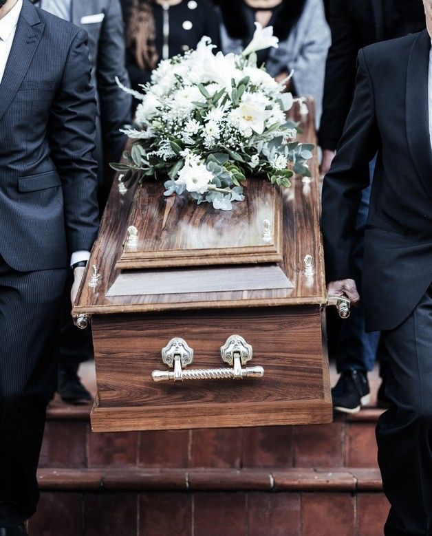 People in suits carrying a wooden casket with white floral arrangement, walking up stairs.