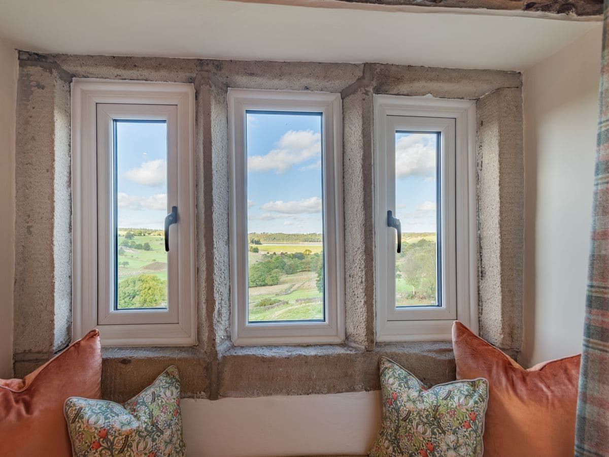 Three white-framed windows offer a view of a green field under a blue sky. Orange pillows sit below.