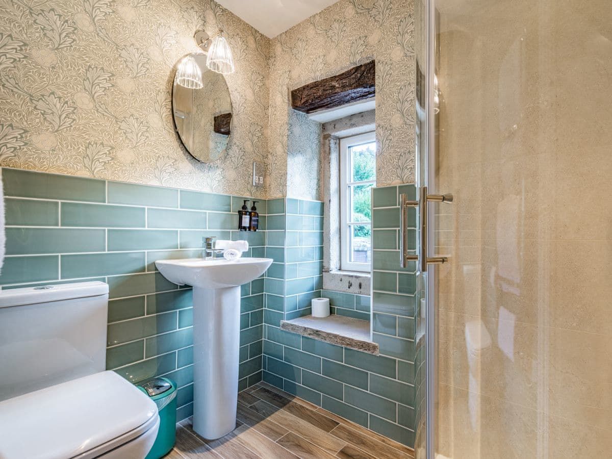 Bathroom with teal tiled walls, white pedestal sink, toilet, and glass shower. Round mirror and small window.