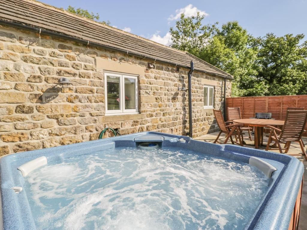 Hot tub in front of a stone cottage on a sunny patio with chairs and table.