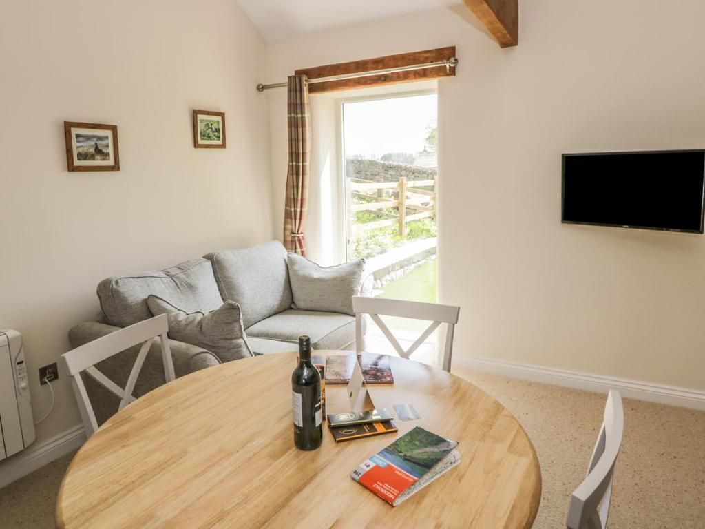Living room with a gray sofa, round wooden table, white chairs, and TV. A window offers a view outside.
