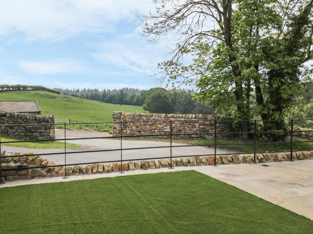 Green field with stone wall, black metal fence, and a paved driveway under a blue sky.