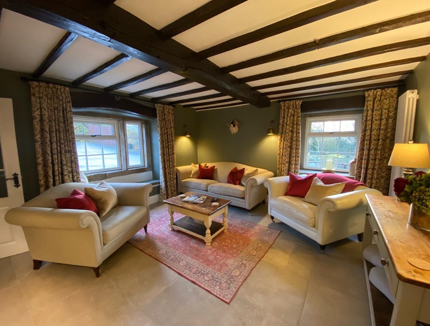 Cozy living room with cream-colored sofas, red pillows, rug, wooden beams, windows, and green walls.