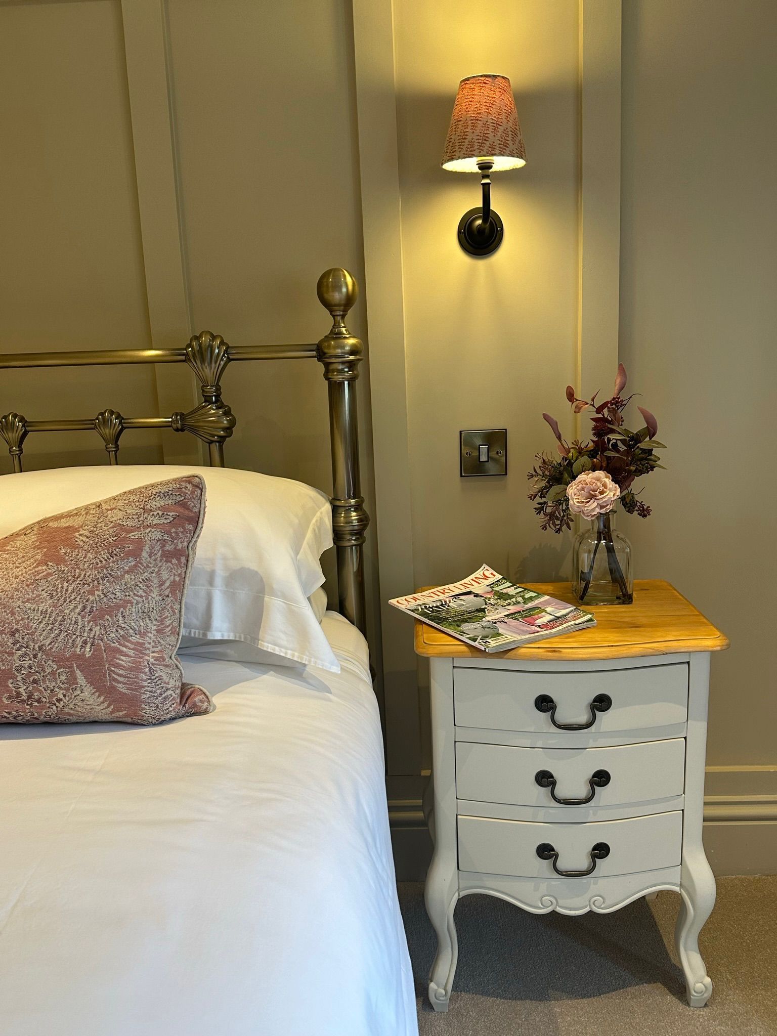 Bedroom interior with bed, nightstand, lamp, and flowers. Light-colored walls and bedding, floral pillow.