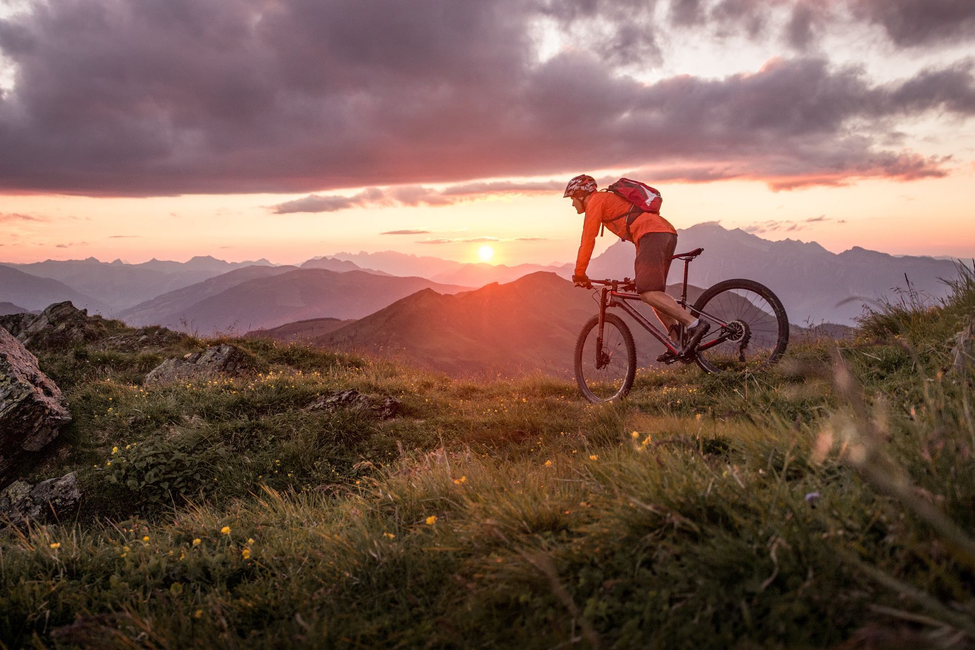 Mountain biker on a trail overlooking mountains at sunset. Rider in orange jacket, against sky's orange and purple hues.