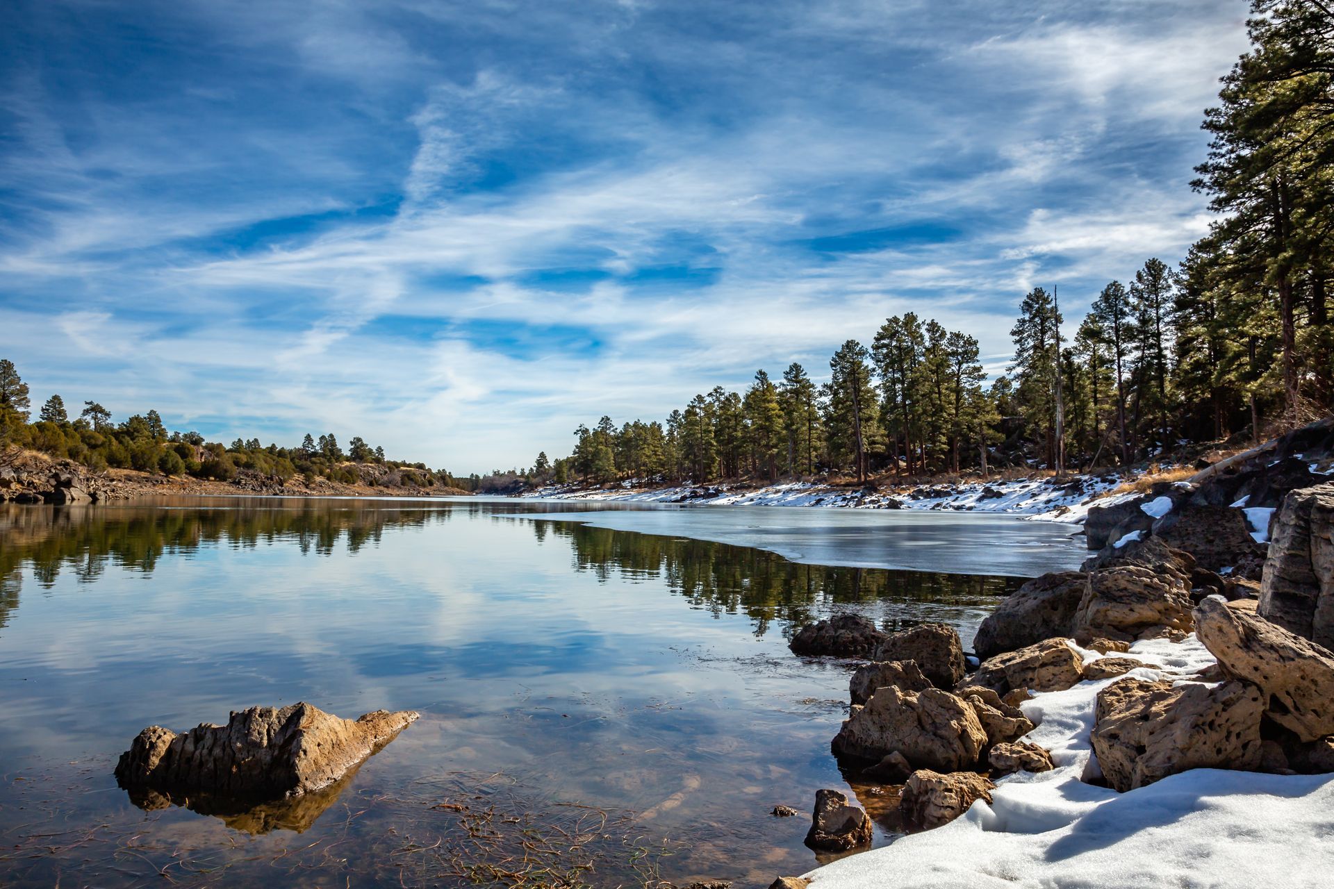 Still lake reflects blue sky, forest, snow patches.