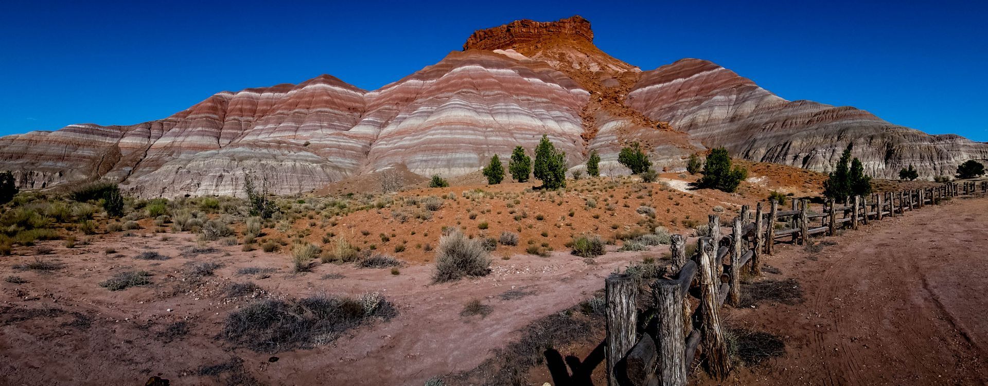 Landscape view of a colorful mountain with layered rock formations and a blue sky. A fence is in the foreground.