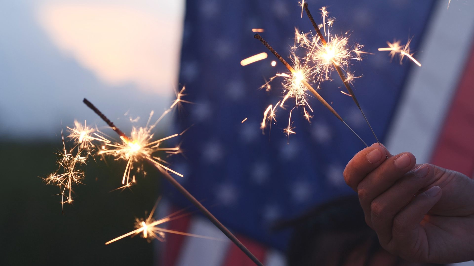Hand holding sparklers against a backdrop of the American flag, glowing in the dusk.